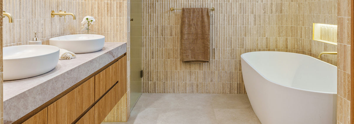Salle de bain travertin – mitigeurs de vasque dorés et baignoire îlot – Valcourt Robinetterie